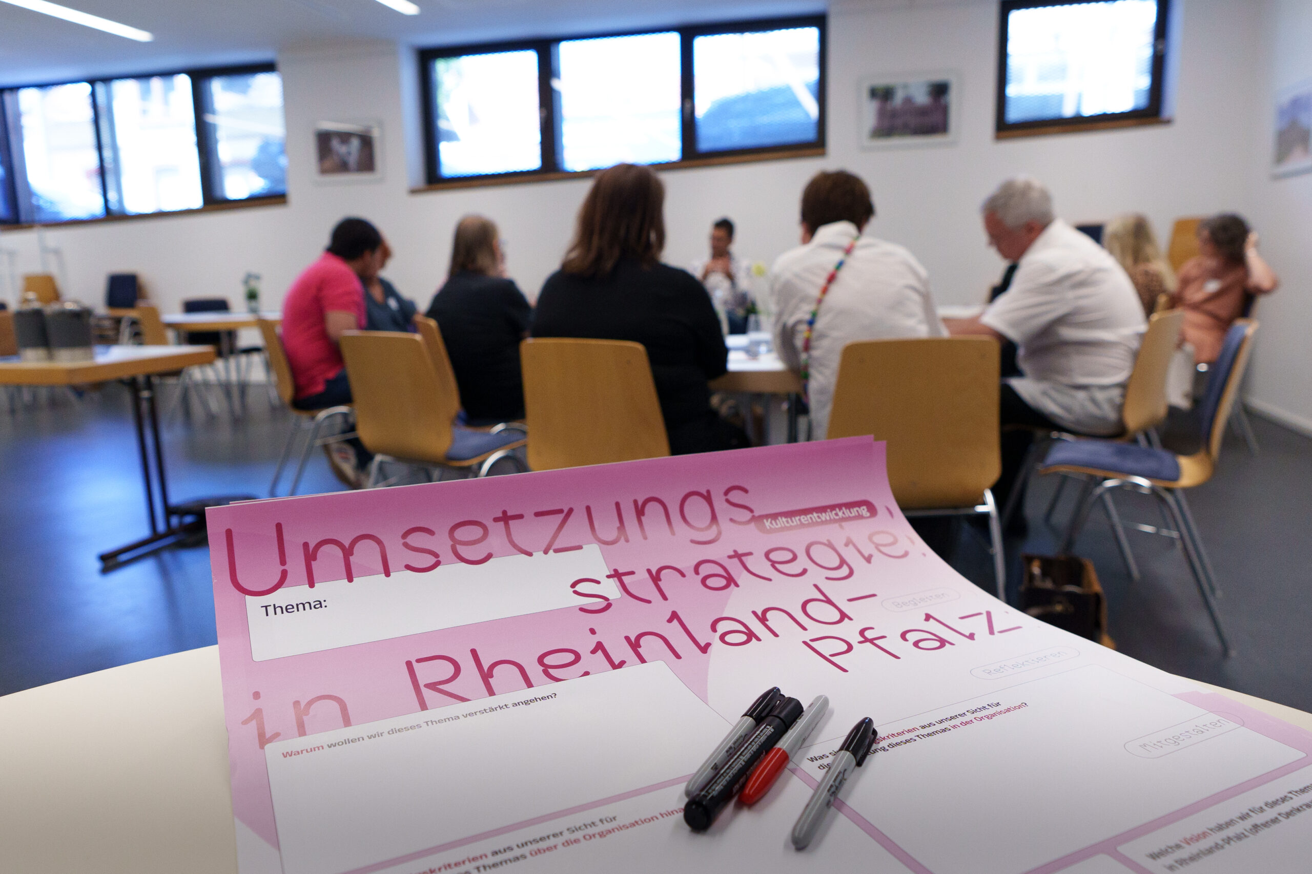 Blick auf eine Diskussionsrunde im Hintergrund. Im Vordergrund ein Plakat mit Stiften. Auf dem Plakat steht Umsetzungsstrategie in Rheinland-Pfalz