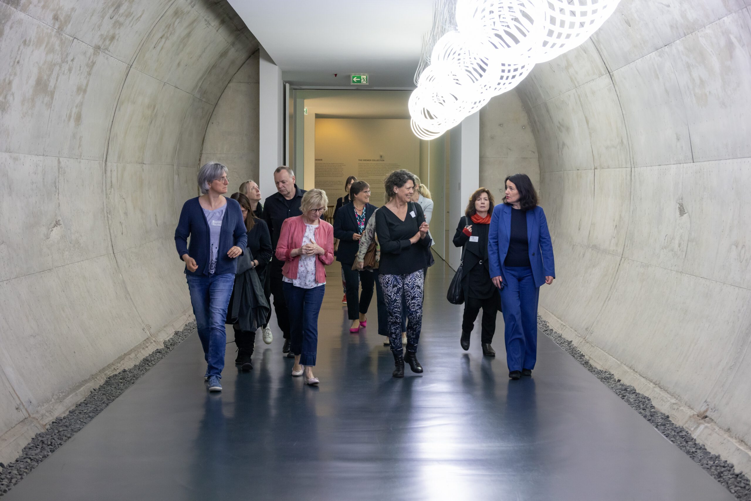Eine Gruppe läuft zu einer Führung im Arp Museum durch einen Tunnel mit einer Lichtinstallation. Rechts ist Frau Dr. Wallner, die Direktorin in einem blauen Anzug zu sehen.