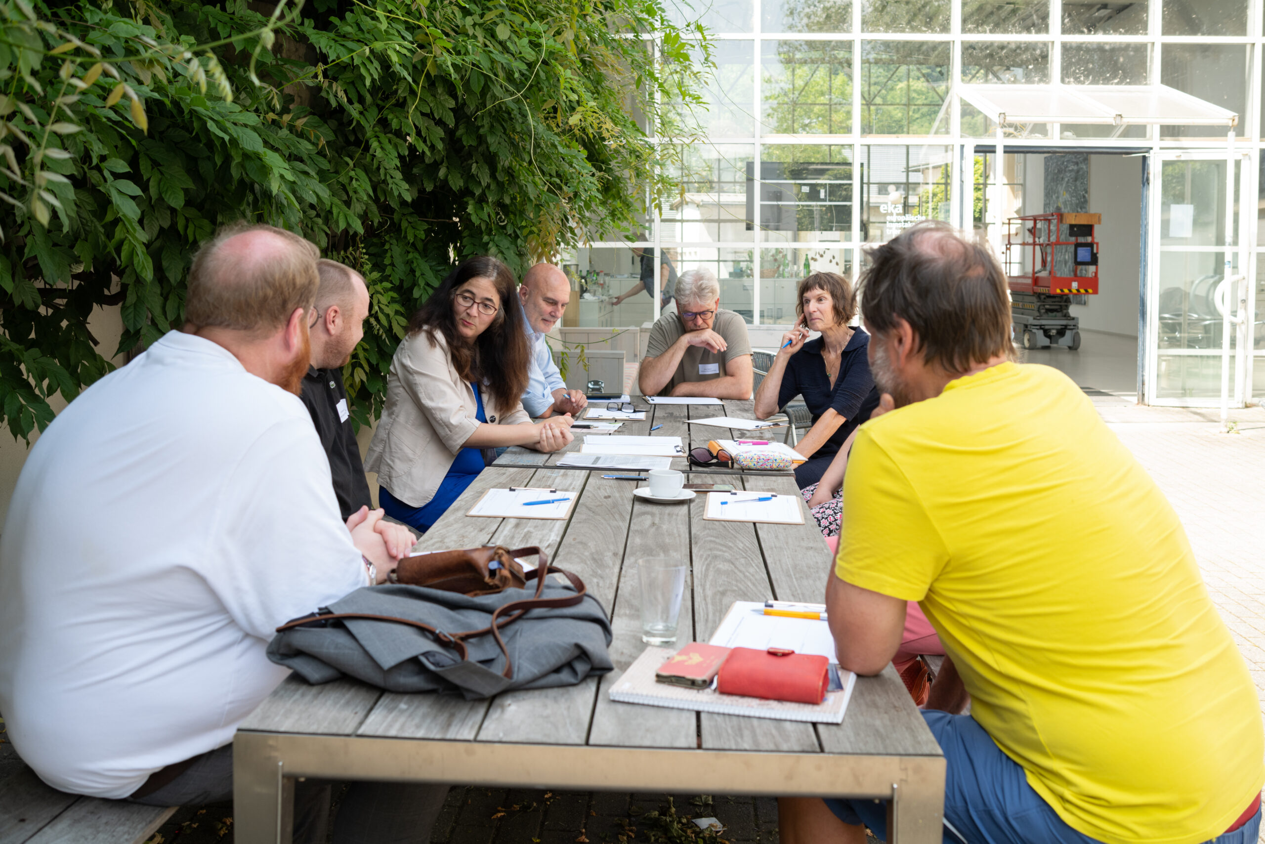 Diskussionsrunde im Freien an einem Holztisch vor grün bepflanzter Wand