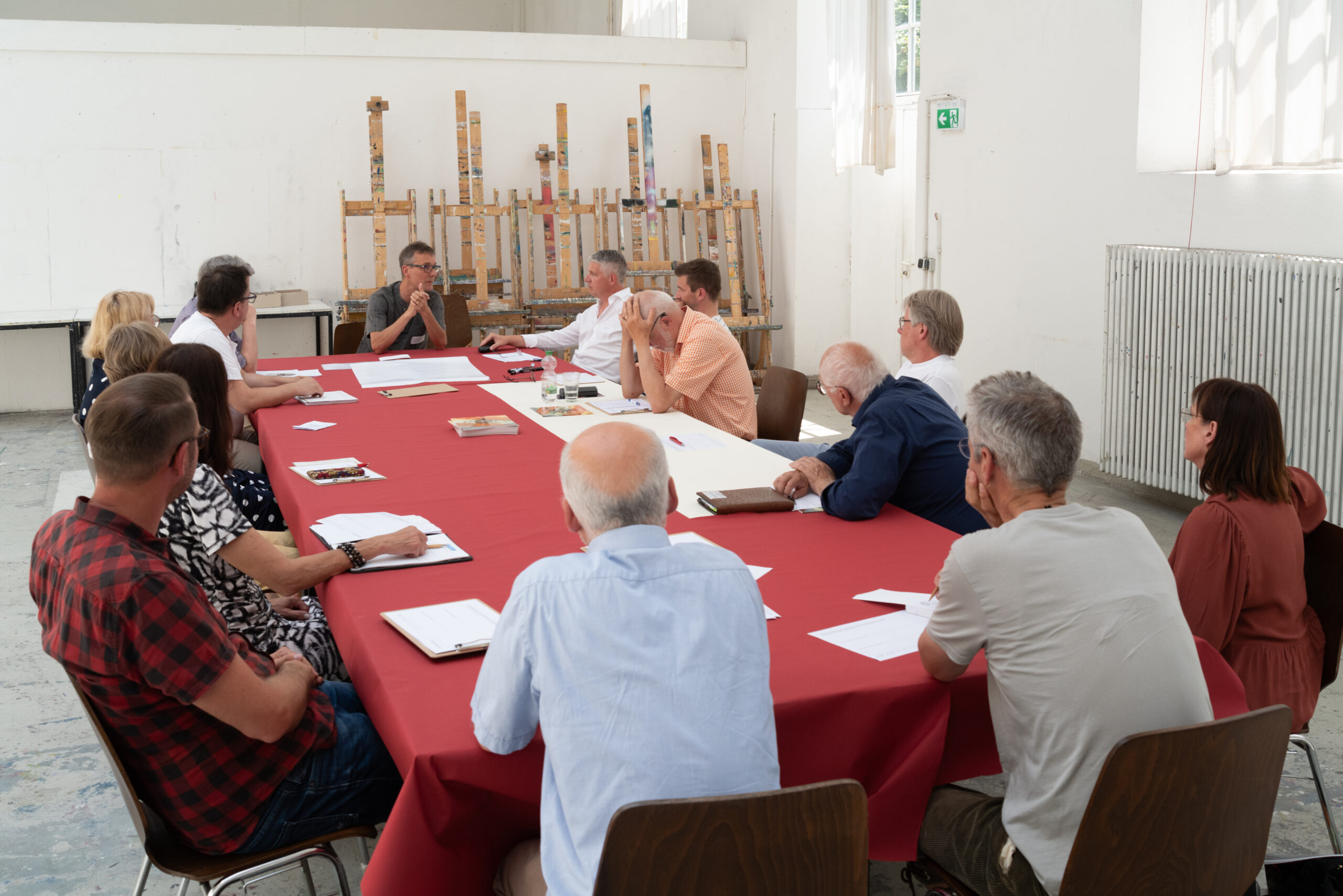Diskussion einer Gruppe in einem Atelier. Die Personen sitzen an einem Tisch mit roter Tischdecke. Im Hintergrund stehen Staffeleien.