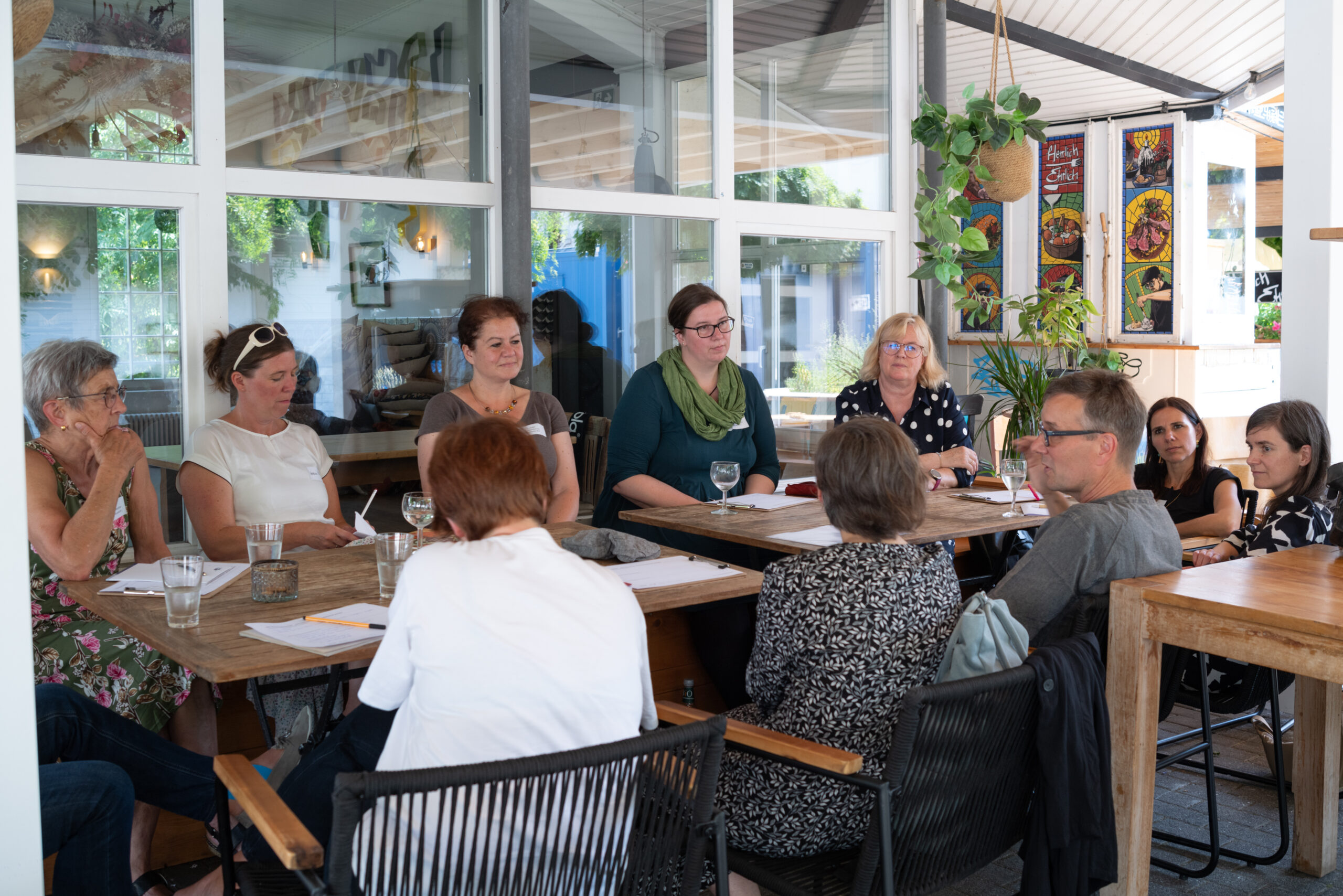 Diskussionsrunde mit 10 Personen, die in einem Café auf einer Außenterrasse an Holztischen sitzen